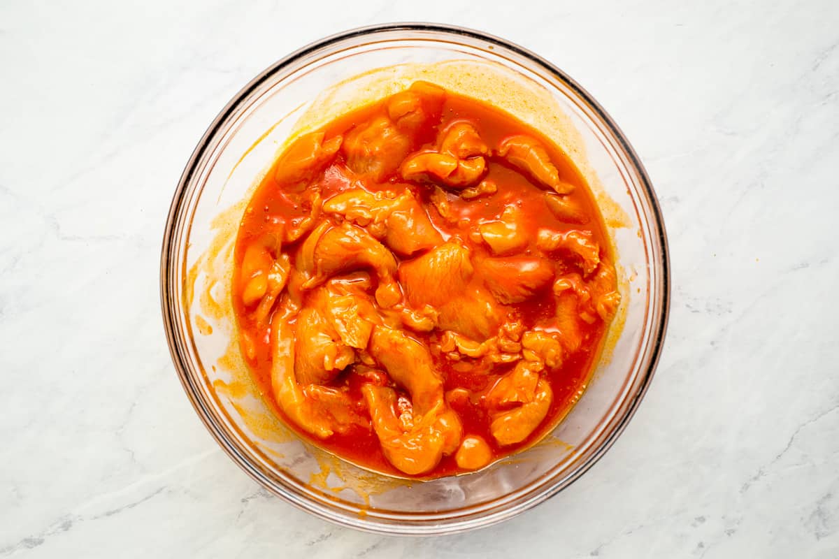 Pieces of chicken marinating in buffalo sauce, in a large glass bowl.