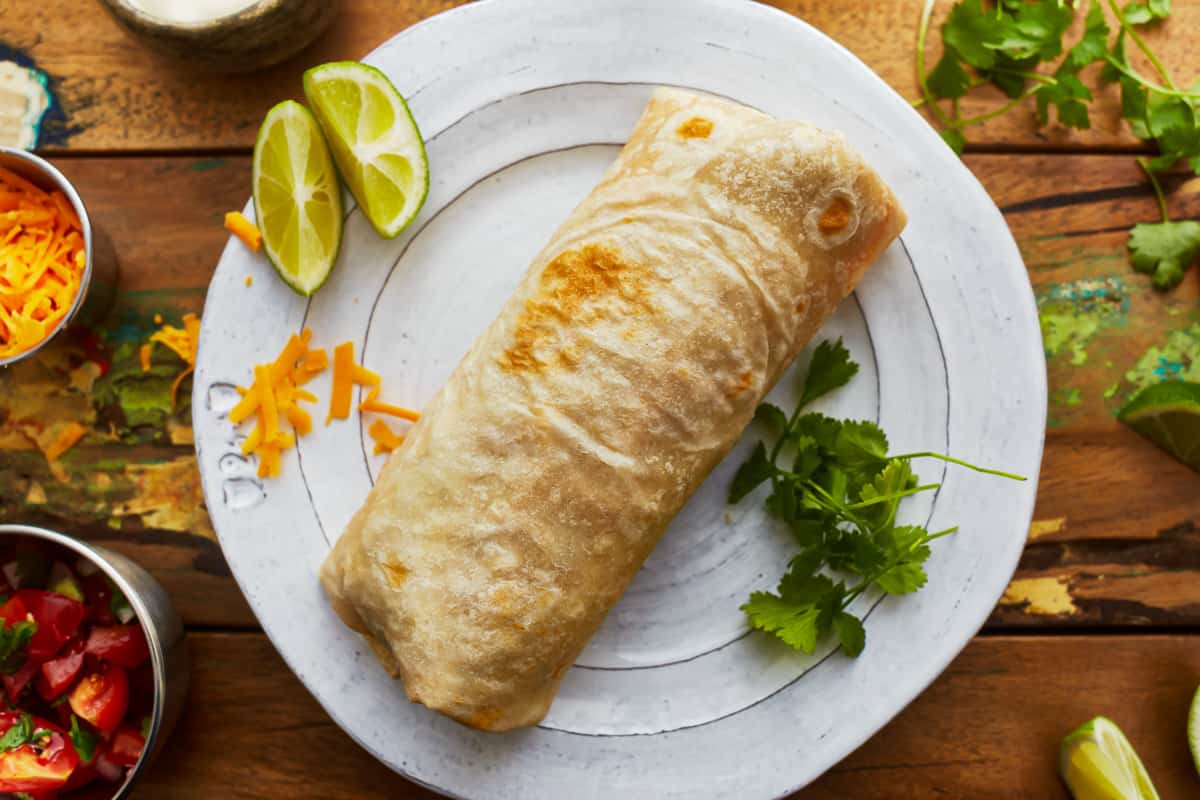 A burrito on a white plate with limes and cilantro.