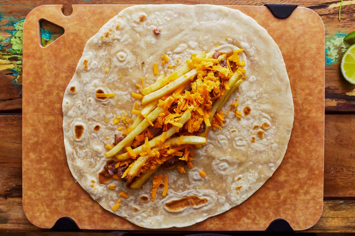 A flour tortilla on a cutting board filled with steak, cheese, and french fries.