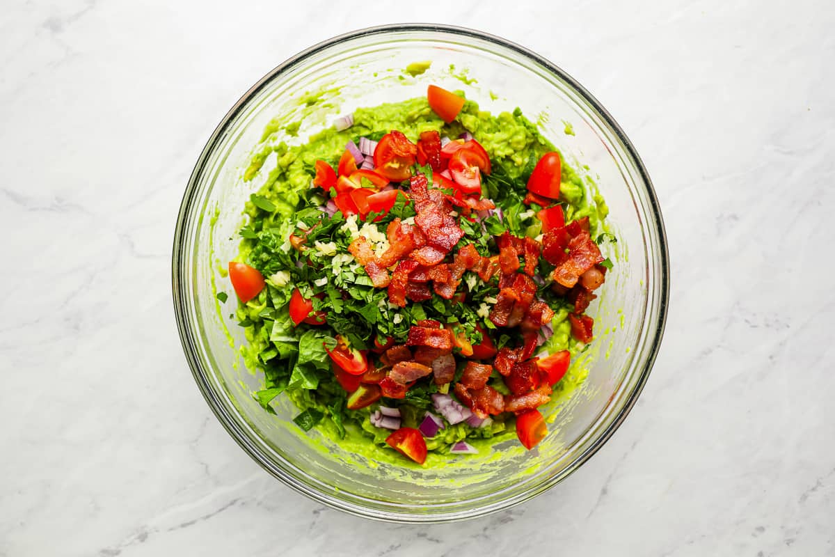 bacon, lettuce, and tomatoes on top of mashed avocado in a glass bowl.
