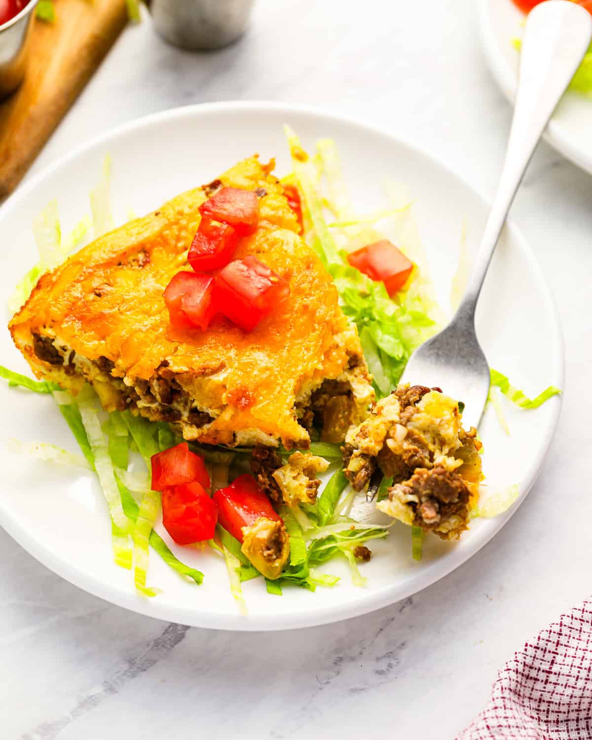 a forkful of cheeseburger pie next to a slice on a bed of lettuce and tomatoes on a white plate.