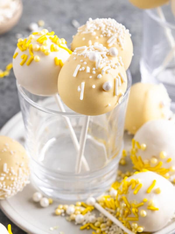 lemon cake pops arranged standing up in a glass, with more cake bites scattered around a plate, with sprinkles and edible pearls as decoration.