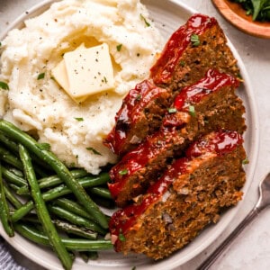 overhead view of 3 slices of meatloaf on a white plate with buttered mashed potatoes and green beans.