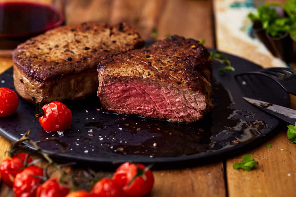 side view of a sliced filet mignon cooked to medium rare on a black plate next to a whole filet.