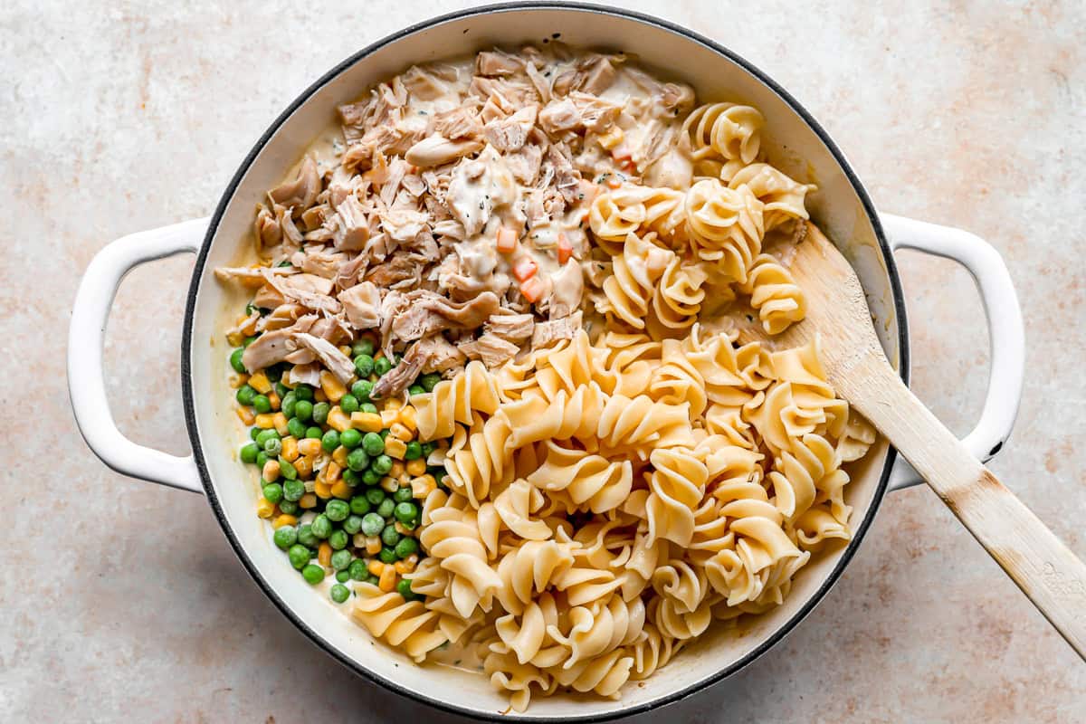 stirring cooked noodles, peas, and shredded chicken into a pot with a wooden spoon.