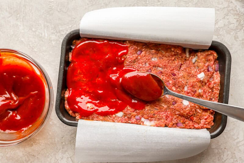 pouring ketchup glaze over meatloaf with a spoon.