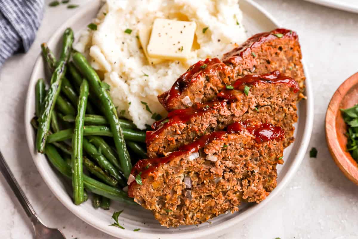 three-quarters view of 3 slices of meatloaf on a white plate with buttered mashed potatoes and green beans.