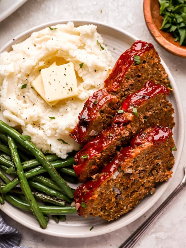 overhead view of 3 slices of meatloaf on a white plate with buttered mashed potatoes and green beans.