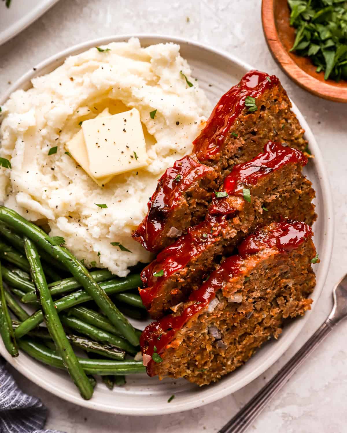 overhead view of 3 slices of meatloaf on a white plate with buttered mashed potatoes and green beans.