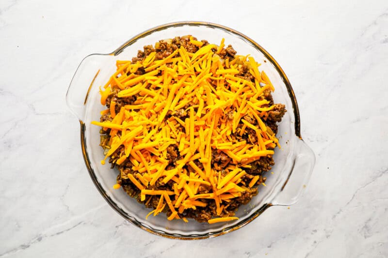 shredded cheese on top of ground beef in a pie pan.