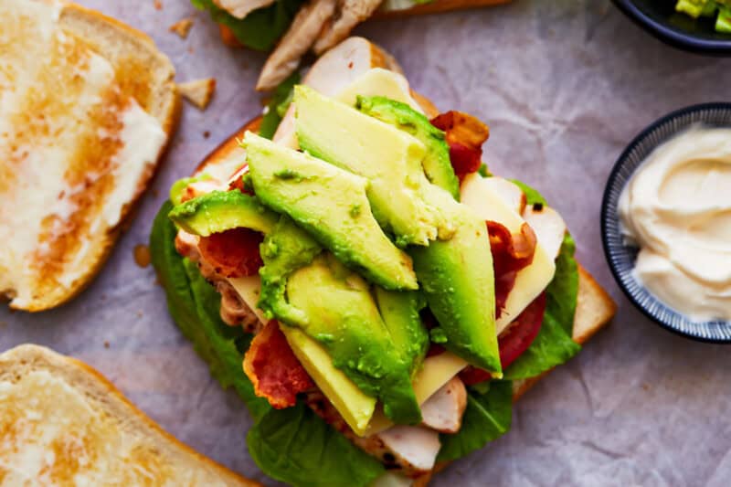 toasted bread topped with lettuce leaves, tomato slices, sliced chicken, cheese, bacon, and avocado.