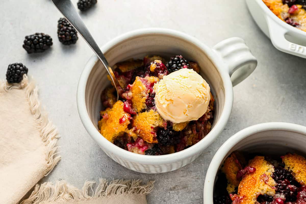 A serving of blackberry cobbler in a small white ramekin, with a scoop of ice cream and a spoon.