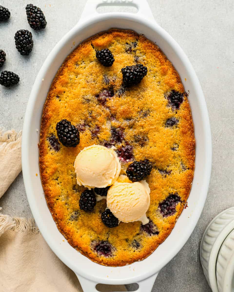 Baked blackberry cobbler in an oval baking dish, topped with ice cream and fresh berries.