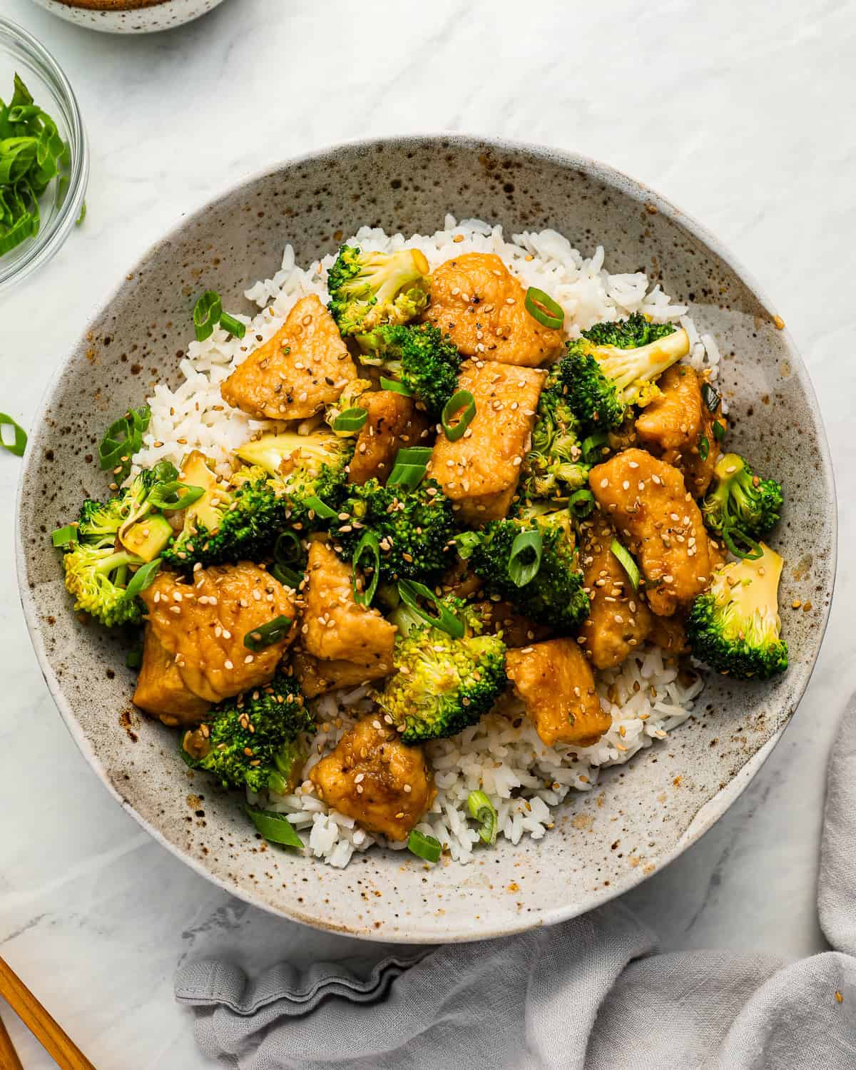 A bowl of chicken and broccoli stir fry served over white rice with green onions and sesame seeds.