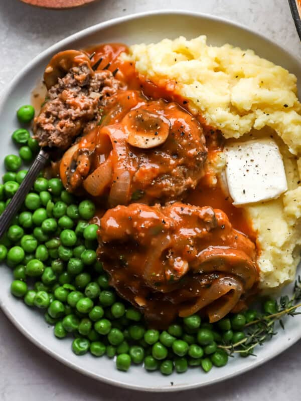 2 hamburger steaks on a white plate with peas, mashed potatoes, and a fork.