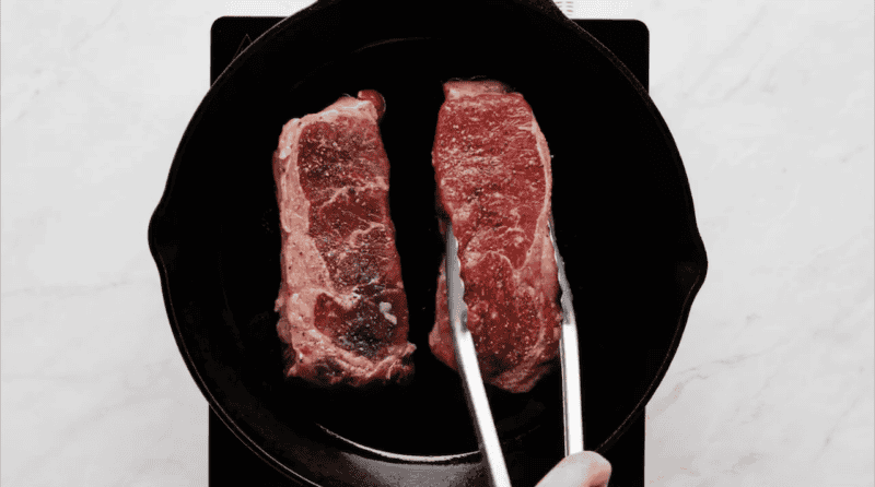 Placing ribeye steaks into a cast iron skillet using metal tongs.
