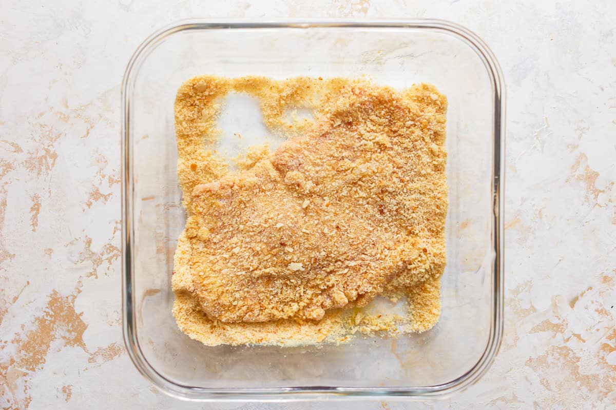 a breaded pork cutlet in a glass bowl filled with breadcrumbs.