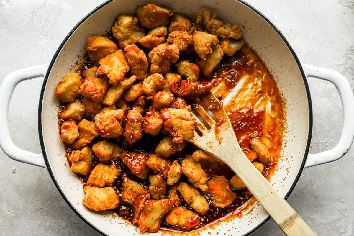 Des morceaux de poulet frits jetés dans une sauce dans une casserole avec une cuillère en bois.