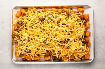 shredded cheese over ground beef and tater tots on a baking sheet.