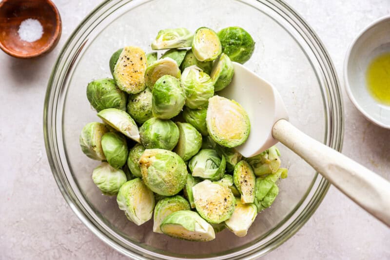 mixing seasoned brussels sprouts in a glass mixing bowl with a rubber spatula.