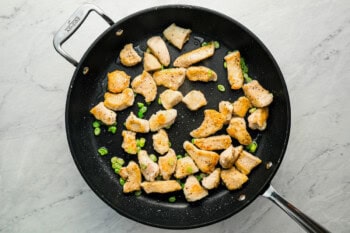 Small pieces of chicken cooking in a skillet with green onions.