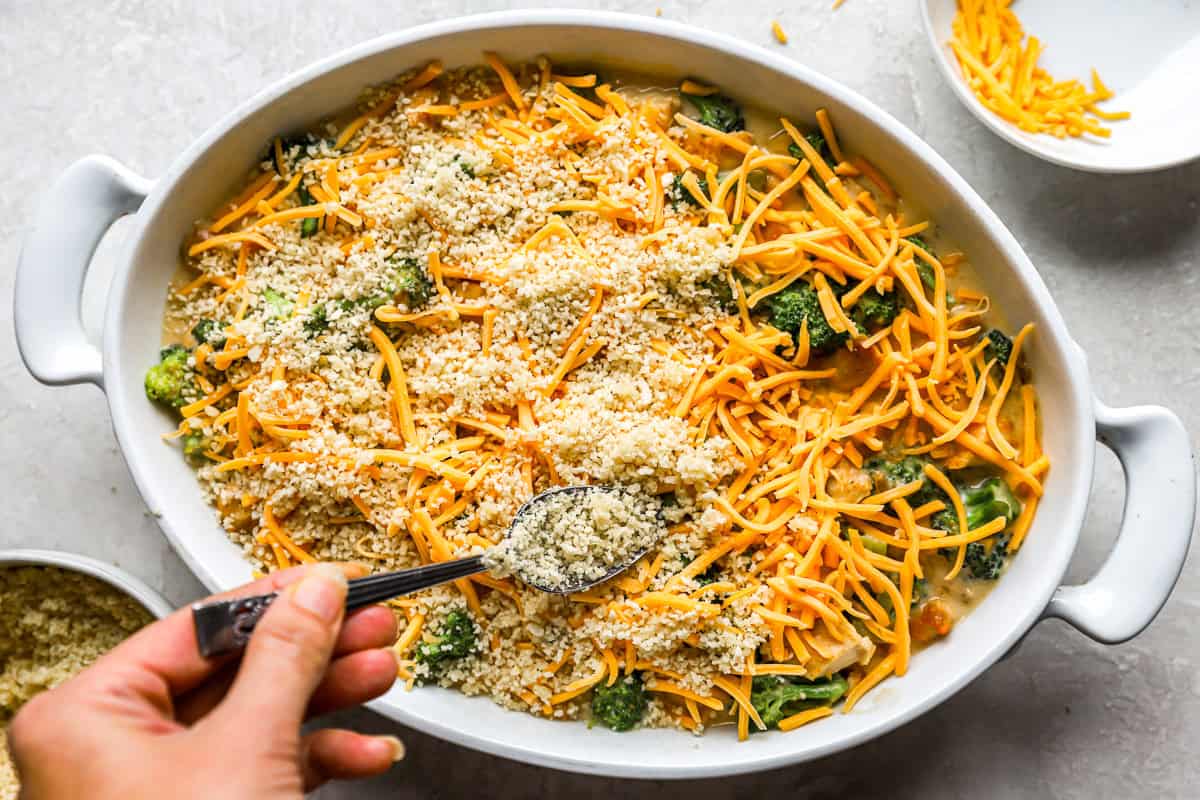 hand holding a spoon, spreading a breadcrumb mixture on top of a casserole.