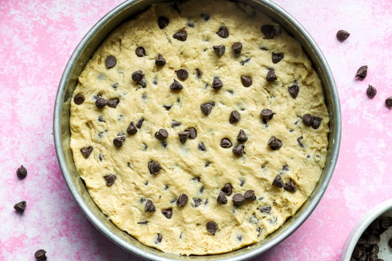 Unbaked cookie cake batter spread into a round cake pan, with chocolate chips sprinkled in.