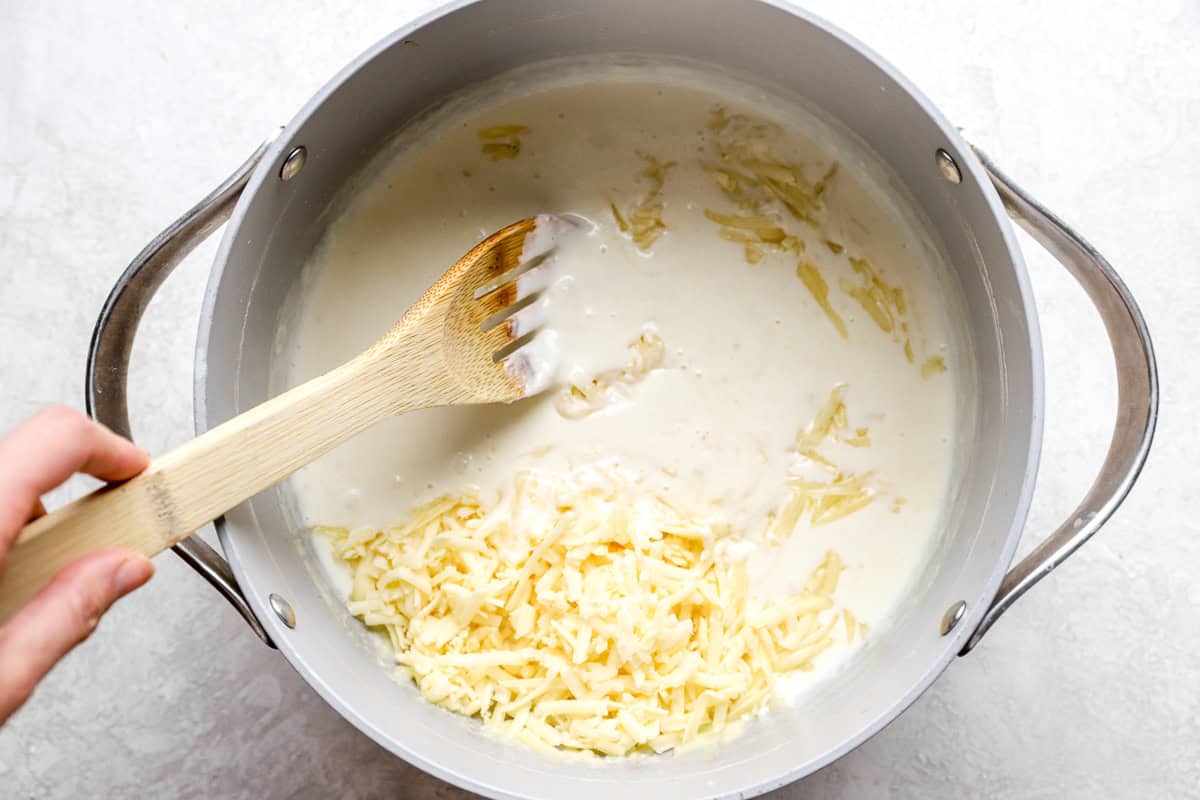 adding cheese to cream sauce in a pot with a wooden slotted spoon.