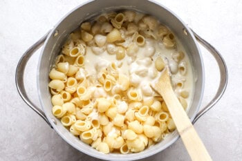 stirring shell pasta into creamy cheese sauce in a pot with a wooden spoon.