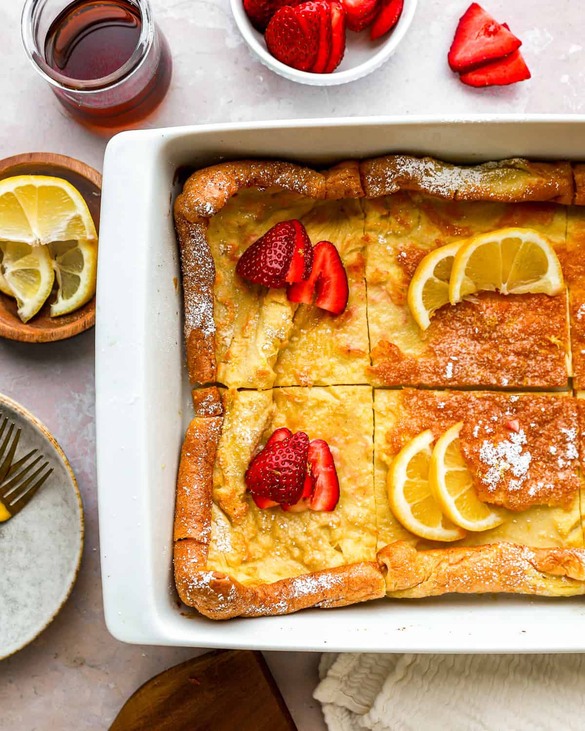 partial overhead view of a cut dutch baby in a rectangular baking pan with lemon slices and strawberries.