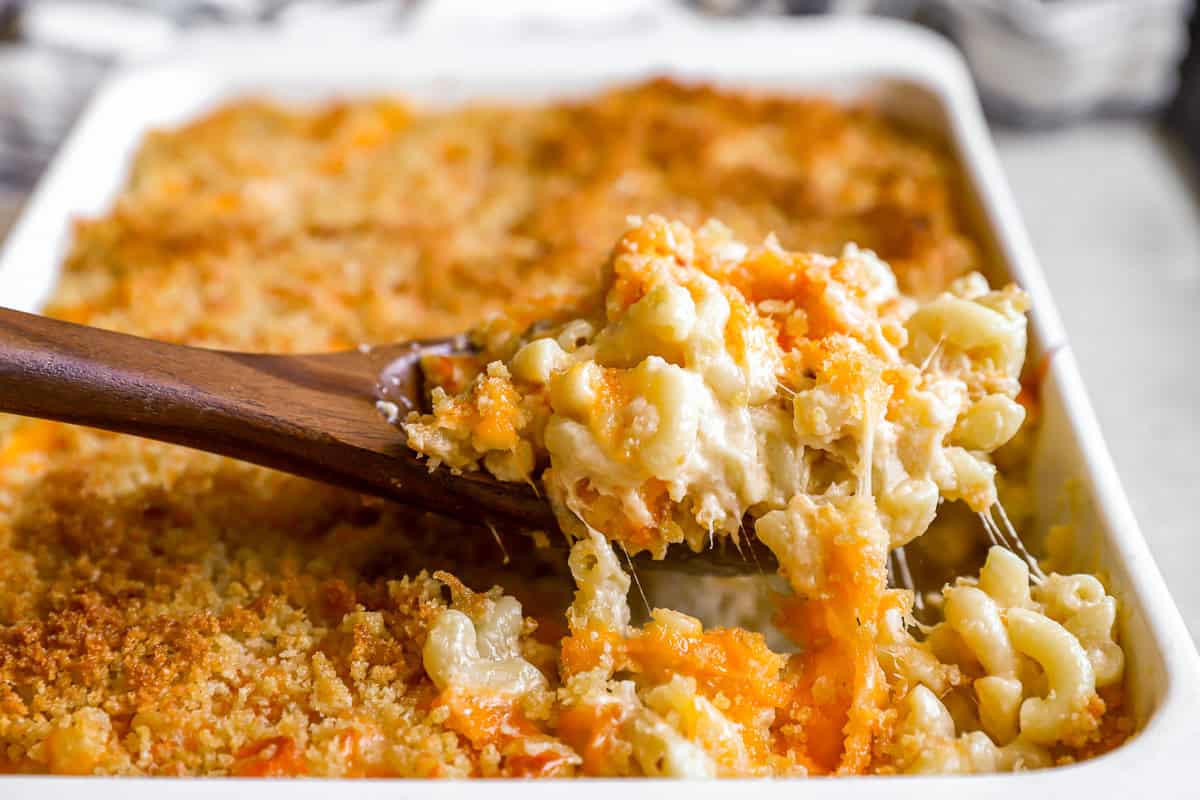 lifting a scoop of baked mac and cheese from a casserole dish with a wooden spoon.