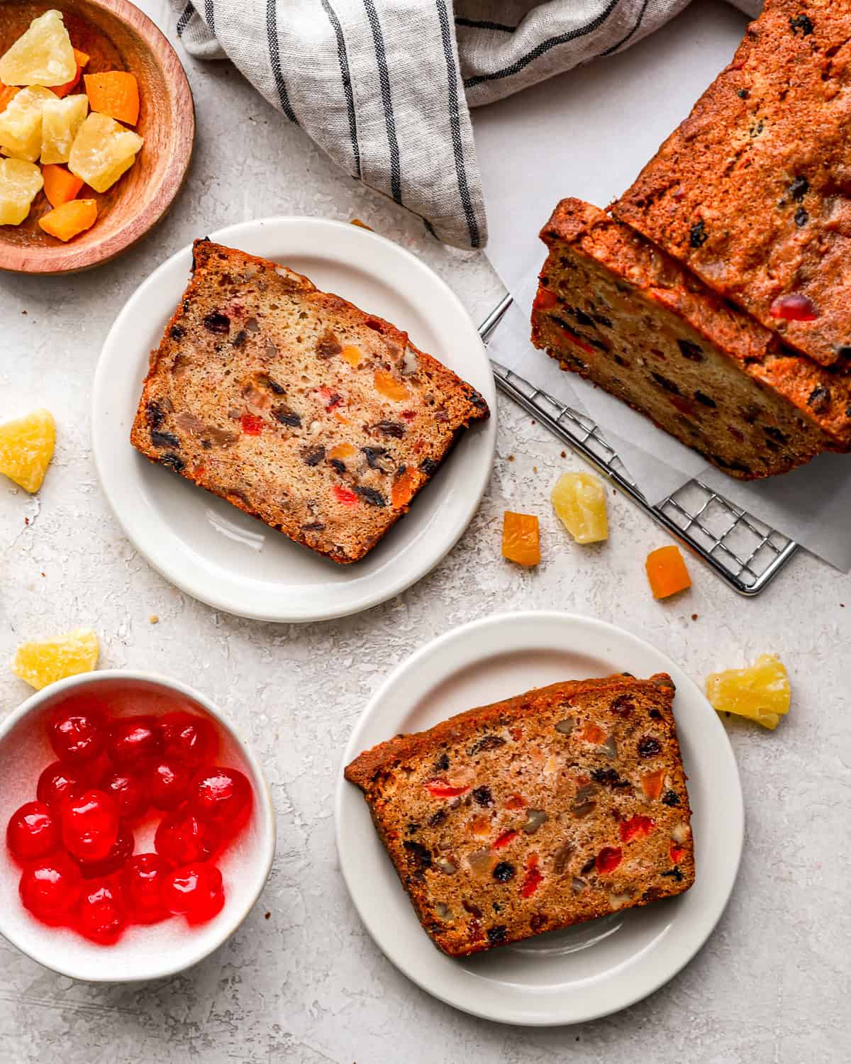 Slices of homemade fruitcake on plates, next to a full loaf and bowls of dried fruits.