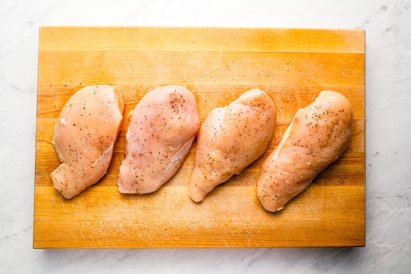 4 seasoned raw chicken breasts on a wooden cutting board.