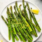 baked asparagus on a white plate with a lemon wedge.