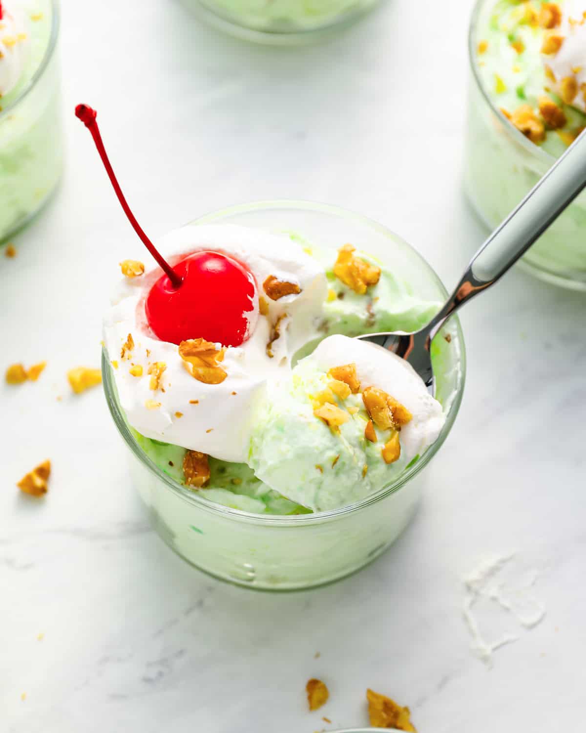 a serving of watergate salad in a glass dish with whipped cream, nuts, a cherry on top, and a spoon.