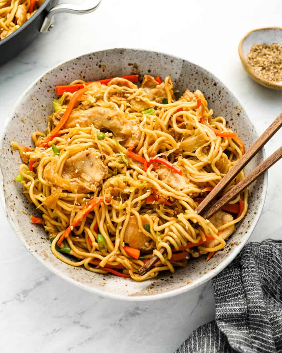 stir fried noodles with chicken and veggies in white specked bowl with chopsticks