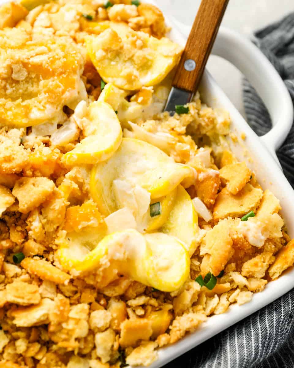 a wooden spoon lifting a scoop of squash casserole from a baking dish.