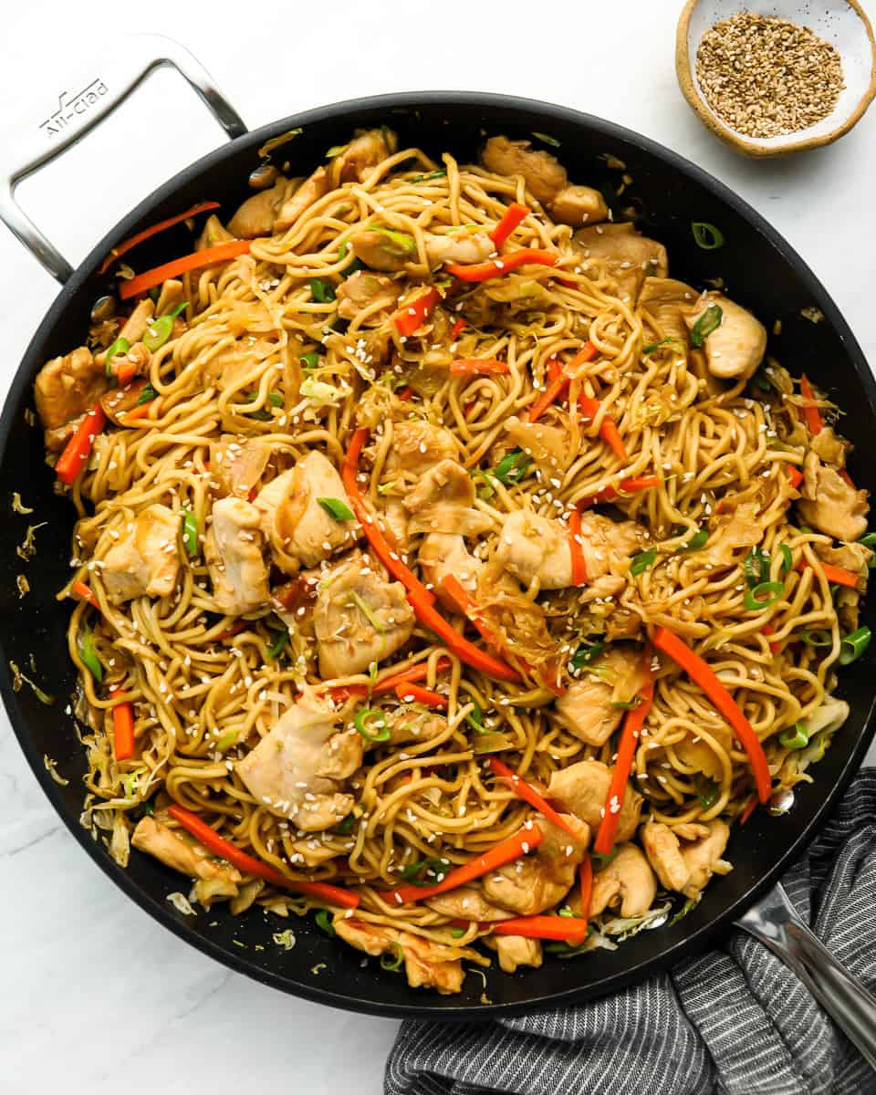A pan filled with stir-fried noodles, chicken pieces, red bell peppers, and green onions, topped with sesame seeds. A small bowl of sesame seeds is placed beside the pan.