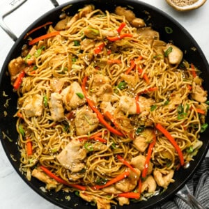 overhead view closeup of stir fried noodles in black pan with chicken, veggies green onions and sesame seeds on top