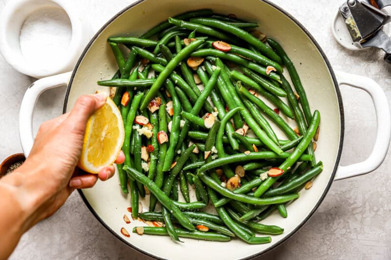 squeezing lemon juice over green beans almondine in a pan.