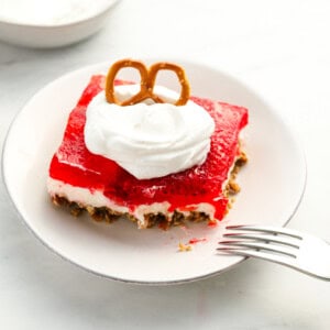 three-quarters view of a partially eaten slice of strawberry pretzel salad on a white plate with whipped cream and a pretzel on top.