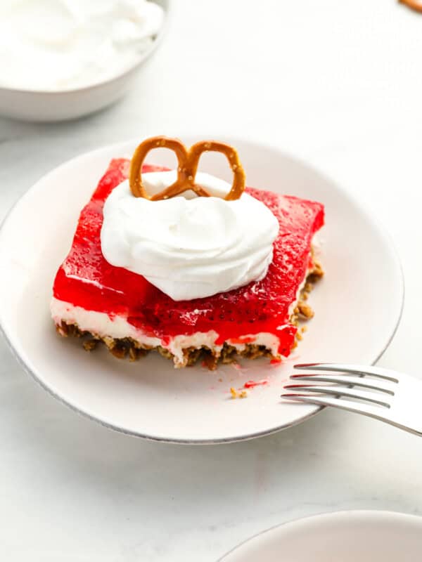 three-quarters view of a partially eaten slice of strawberry pretzel salad on a white plate with whipped cream and a pretzel on top.