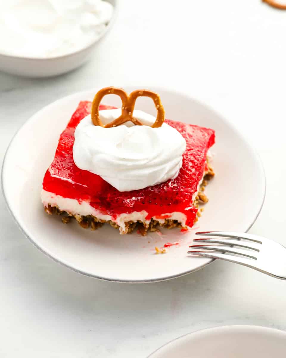 three-quarters view of a partially eaten slice of strawberry pretzel salad on a white plate with whipped cream and a pretzel on top.