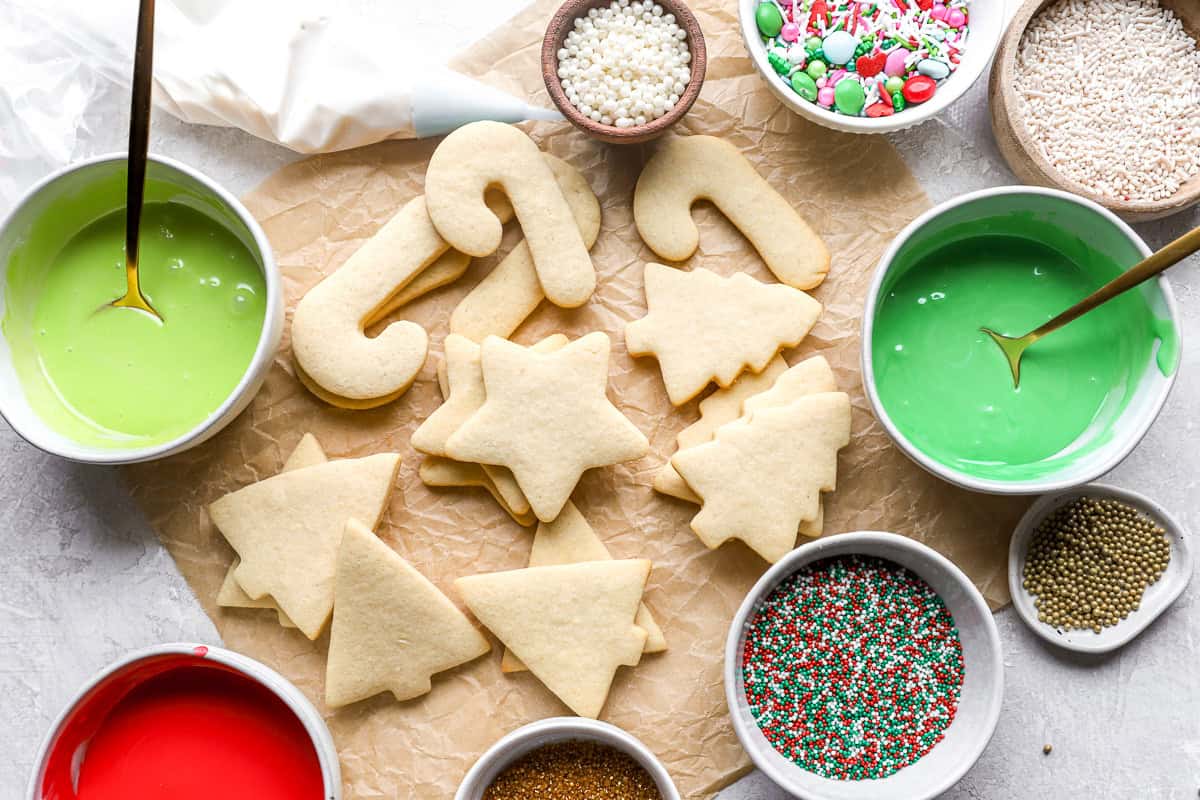 stacked cutout christmas cookies on parchment surrounded by bowls of colored icing and sprinkles.