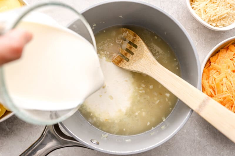 pouring cream over sautéed onions in a pan with a wooden spoon.