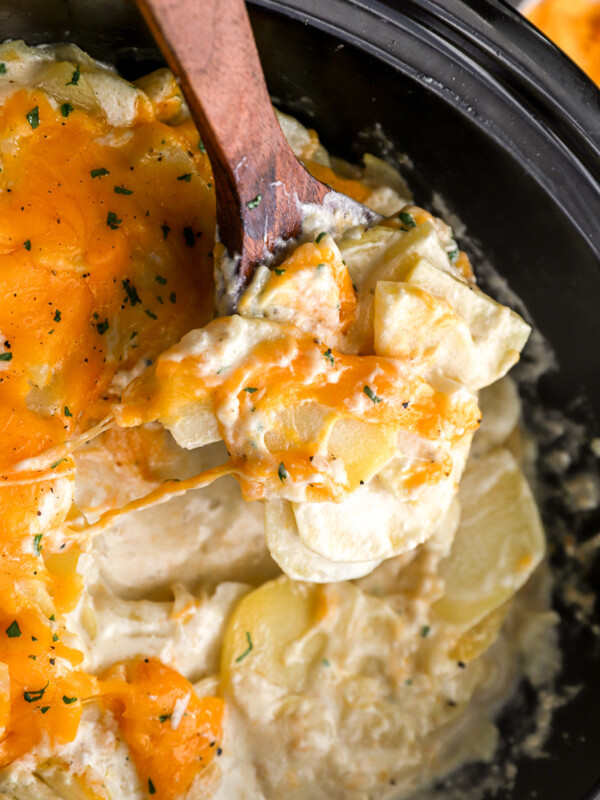 a wooden spoon lifting cheesy scalloped potatoes from a crockpot.