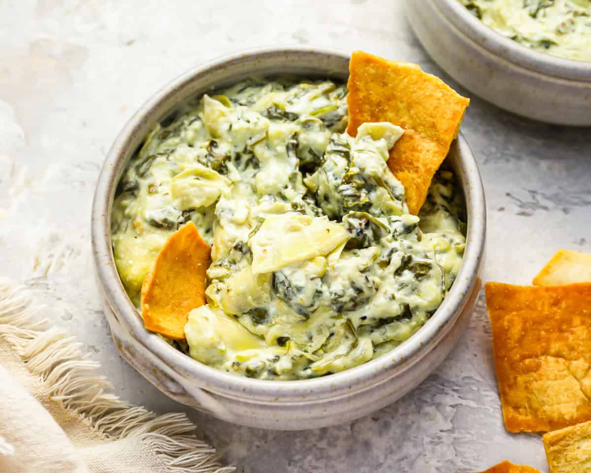 a serving of crockpot spinach artichoke dip in a white bowl with pita chips.