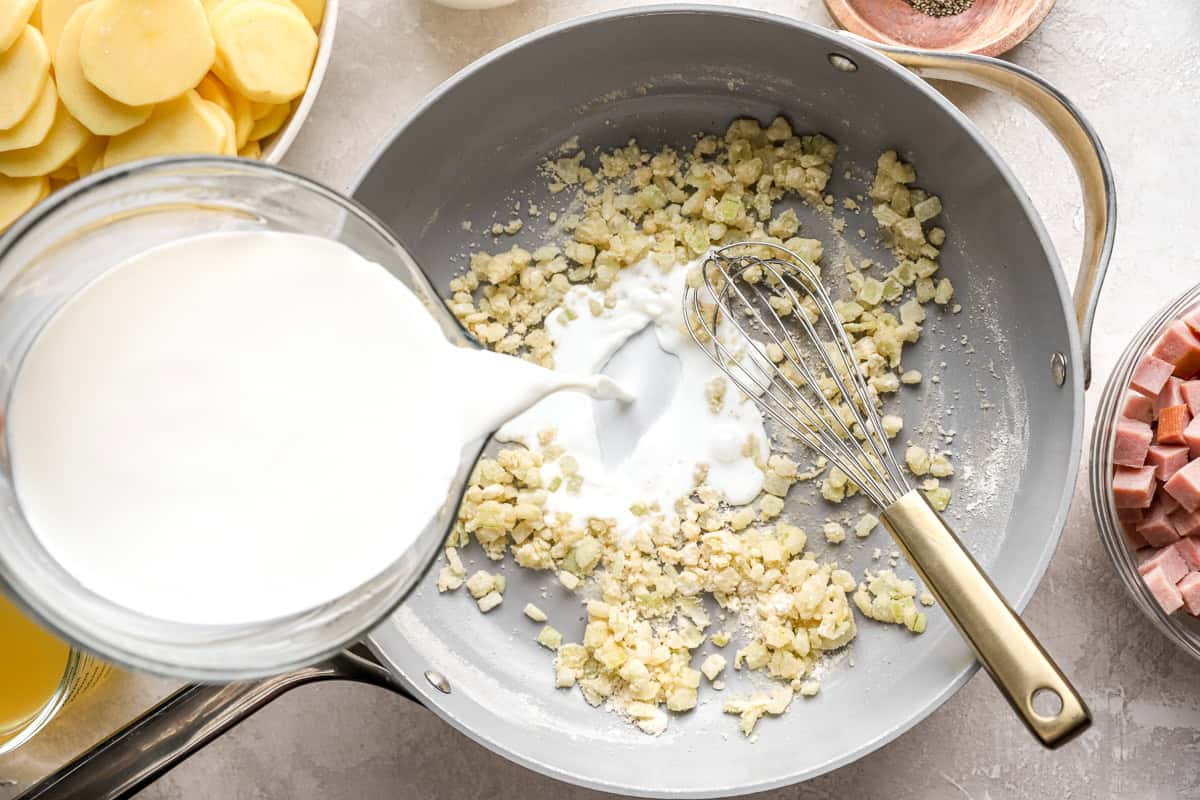 pouring milk over sautéed onion and flour in a pan with a whisk.