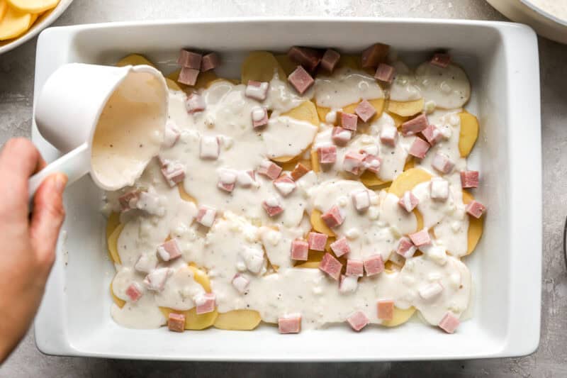 pouring white sauce over thinly-sliced potatoes and ham in a casserole dish.