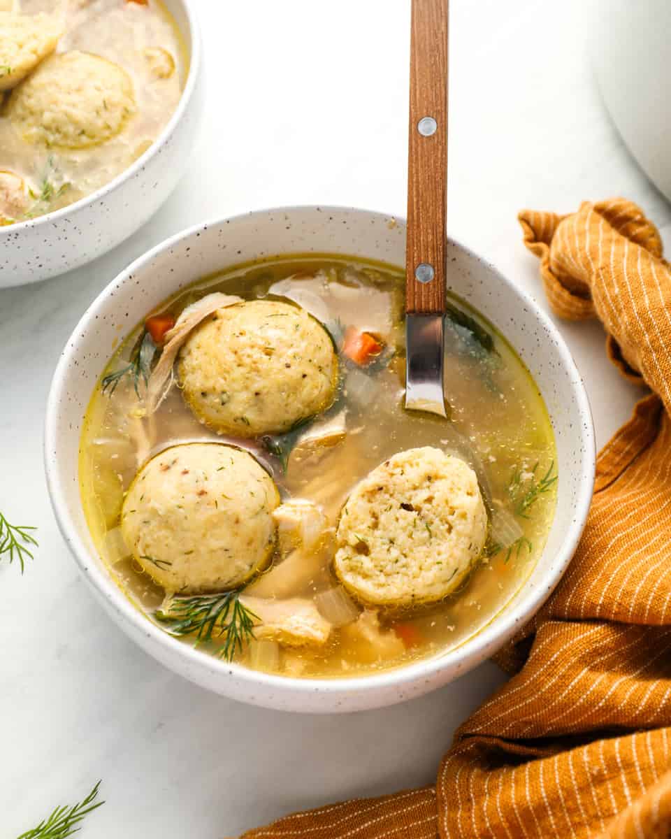 A bowl of matzo ball soup featuring golden matzo balls in a clear chicken broth with vegetables and fresh dill, placed on a white surface with an orange-striped napkin.